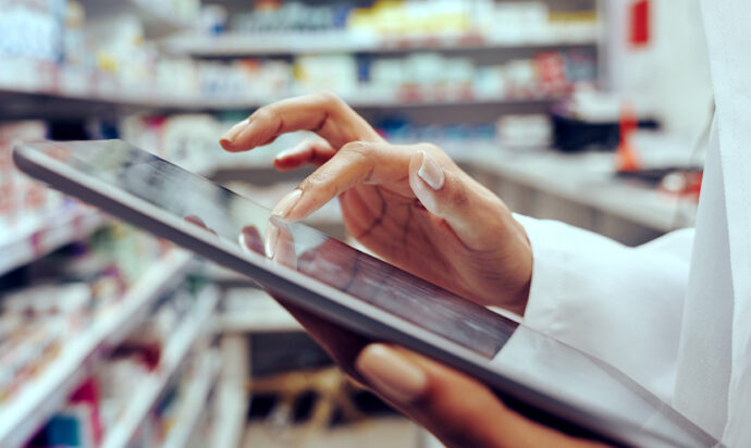 Person in lab coat touching tablet, pharmaceutical ingredients on shelves in background